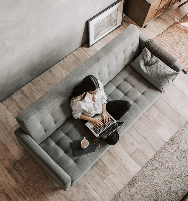 A woman sits cross-legged on a gray sofa, working on a laptop. A mug, notebook, and glasses are beside her. The stylish room features wooden floors—an ideal workspace for a Mortgage Broker Fremantle with a framed picture on the floor.