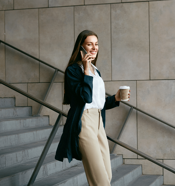 A woman with long brown hair walks down stairs in front of a stone wall, smiling while talking on her smartphone and holding a takeaway coffee cup. She wears a white blouse, beige pants, and a dark jacket—perfect for a busy Mortgage Broker Perth day.