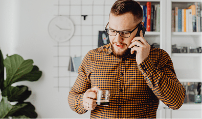 A man wearing glasses and a checkered shirt holds a mug and talks on a smartphone in a modern home office, suggesting the professional workspace of a Mortgage Broker Perth, with bookshelves and a wall clock in the background.